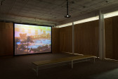A makeshift screening room enclosed by unpainted wooden walls. There are two wooden benches arranged to face a projector screen. The screen displays an image of a luxury pool surrounded by palm trees, with the sun shining.