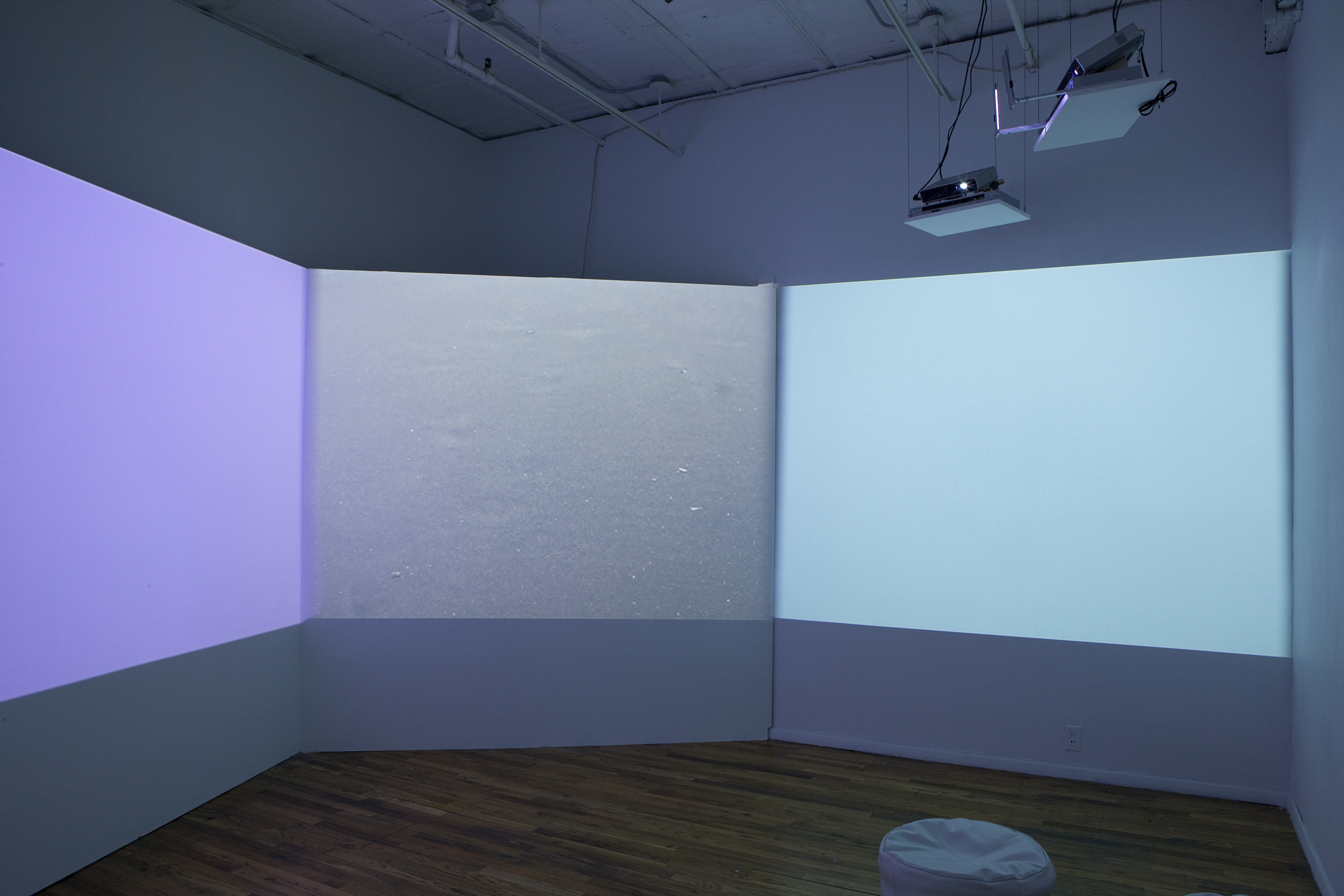 A dimmed room with three projections casting purple, gray, and aqua light against the walls. In the corner of the image is a small, circular seat.