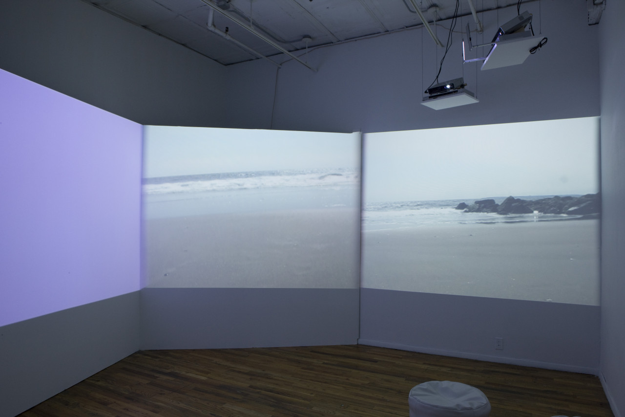 A dimmed room with three projections, one casting purple light, the other two depicting stills of shorelines. In the corner, a small, round seat is visible.