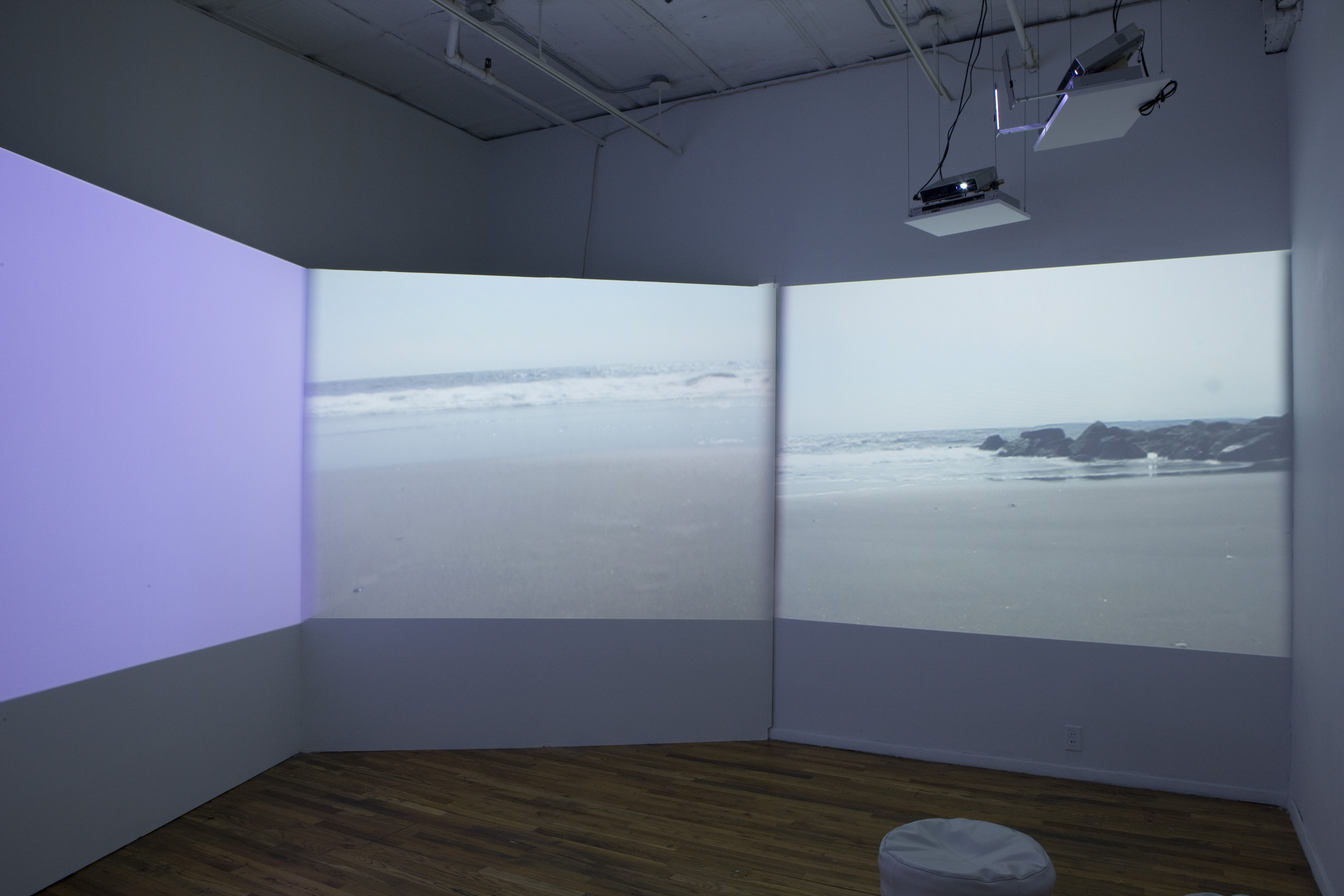 A dimmed room with three projections, one casting purple light, the other two depicting stills of shorelines. In the corner, a small, round seat is visible.