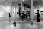 A view of a gallery space with concrete floors, white walls, and white columns.  In the foreground, a grouping of five striped cylinders of varying sizes are placed on the floor.  Other works are visible in the background.