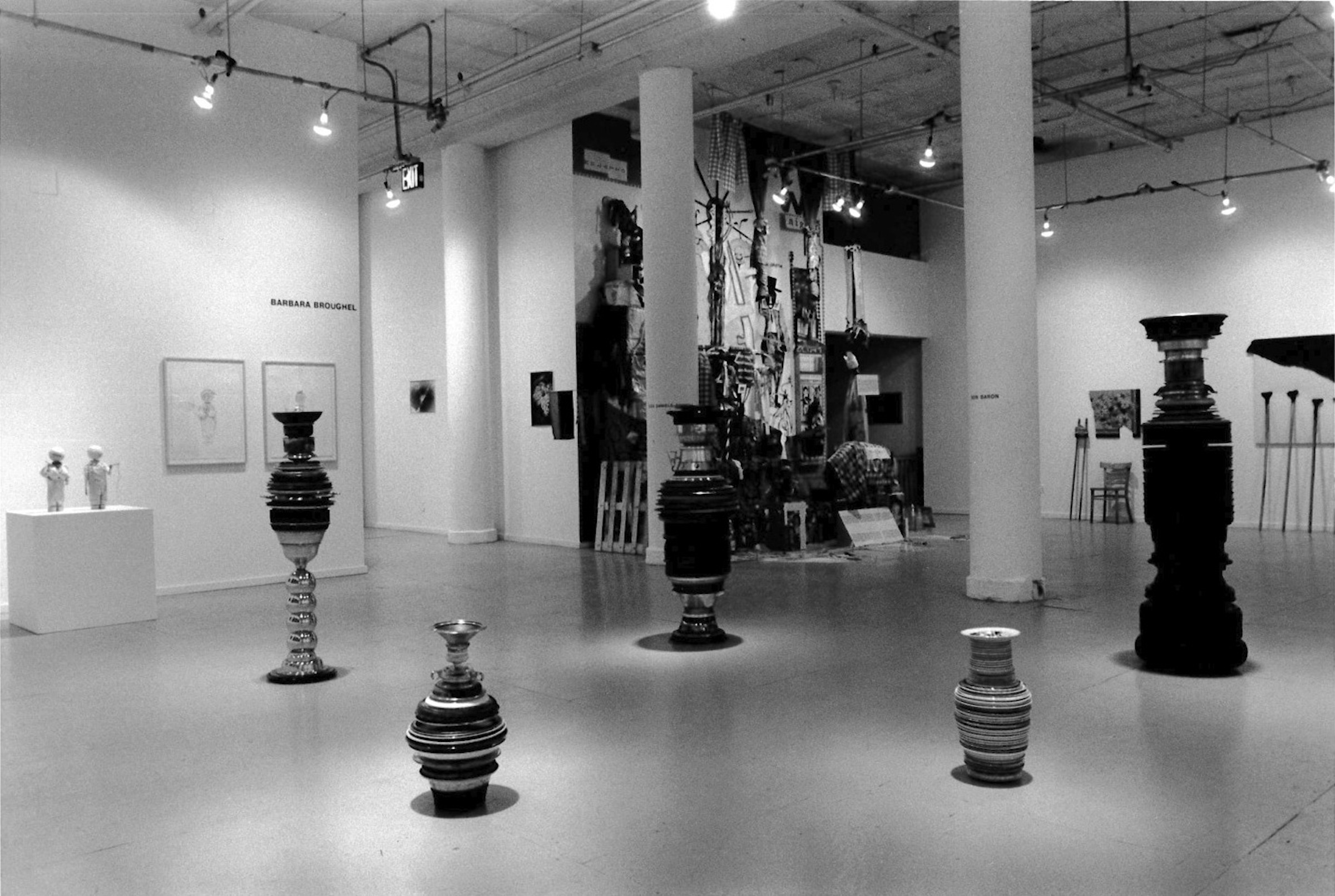 A view of a gallery space with concrete floors, white walls, and white columns.  In the foreground, a grouping of five striped cylinders of varying sizes are placed on the floor.  Other works are visible in the background.