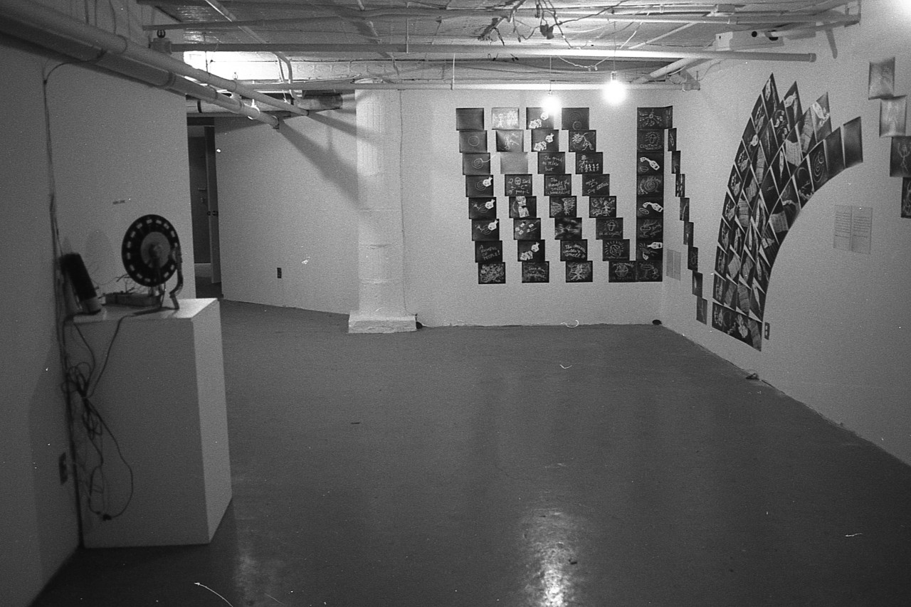 A photograph of a low-ceilinged gallery space.  On the right, drawings on black paper are arranged in grids and arches on the wall.  On the left, electrical equiment sits on top of a pedestal.
