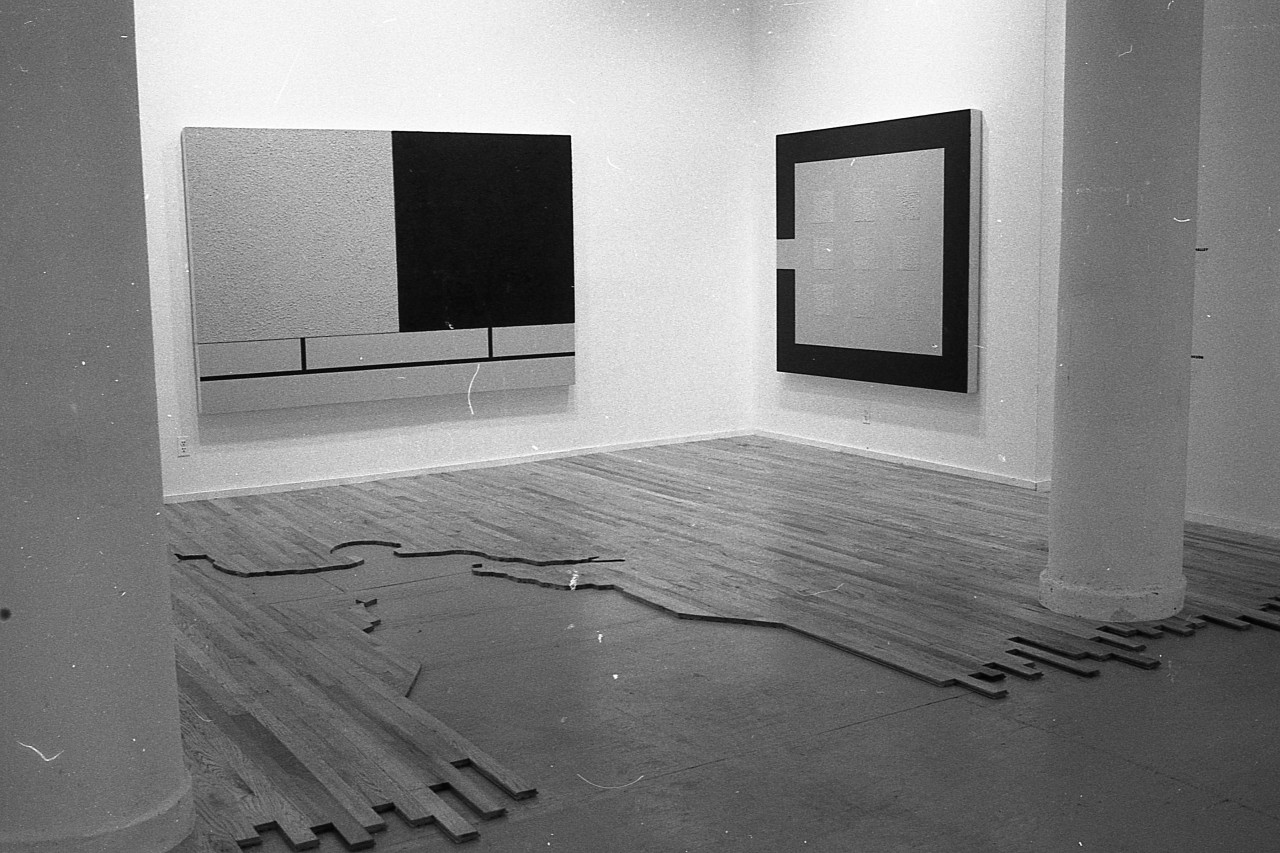 A photograph of a large gallery space with multiple rooms visible.  In the foreground, the floor is covered with flooring, except for a silhouette of a human figure which has been cut out of the flooring, through which the concrete below is visible.  Two geometric abstract paintings hang on the wall above.