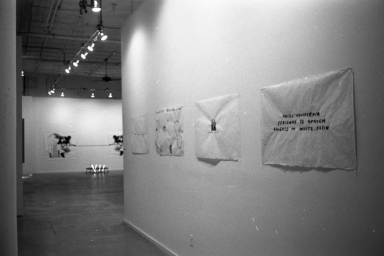 A photograph showing a row of text-based paintings hanging on a gallery wall.  Beyond them, another gallery space with other artworks is visible.