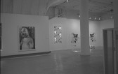 A photograph of paintings hanging on the walls of a large gallery space with white walls.  The work closest to the camera is a realistic painting of a baby, shown for the waist up.
