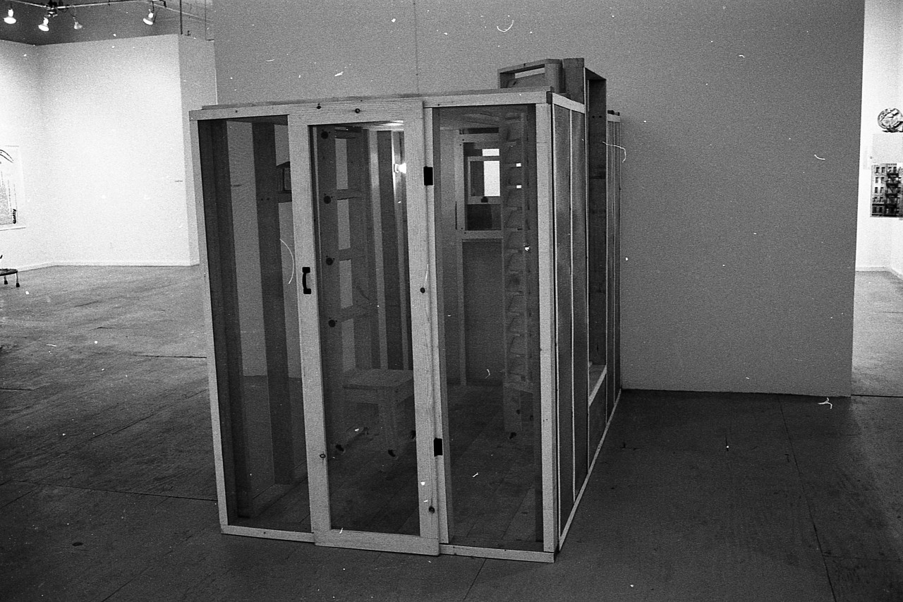 A photograph of a small room with clear walls and a clear door sitting against a wall in a white-walled gallery.  Inside the room, a wooden chair and shelves are visible.