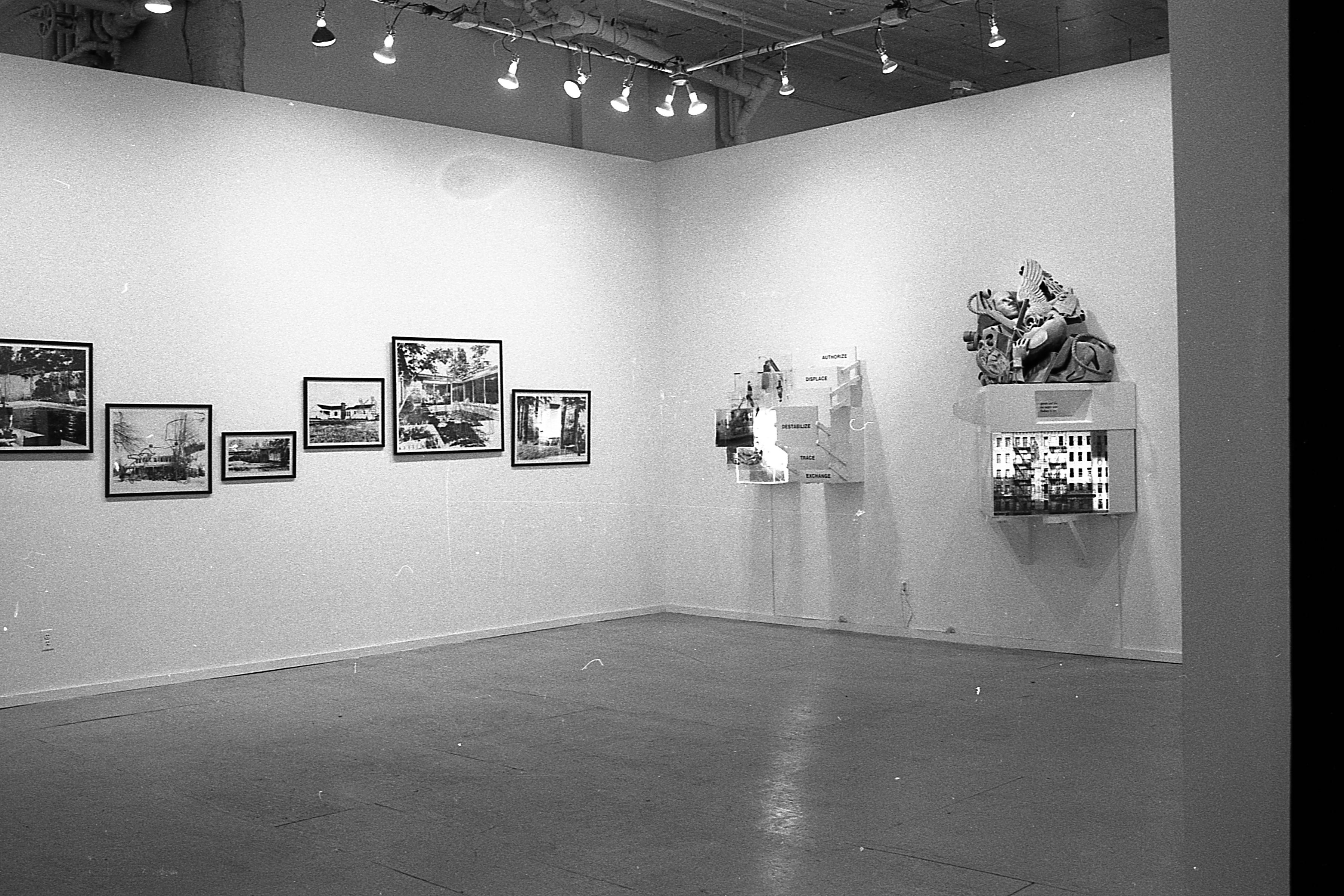A photograph of two walls of a gallery space.  On one, photographs hang in an uneven row.  On the other, two sculptures protrude from the wall.