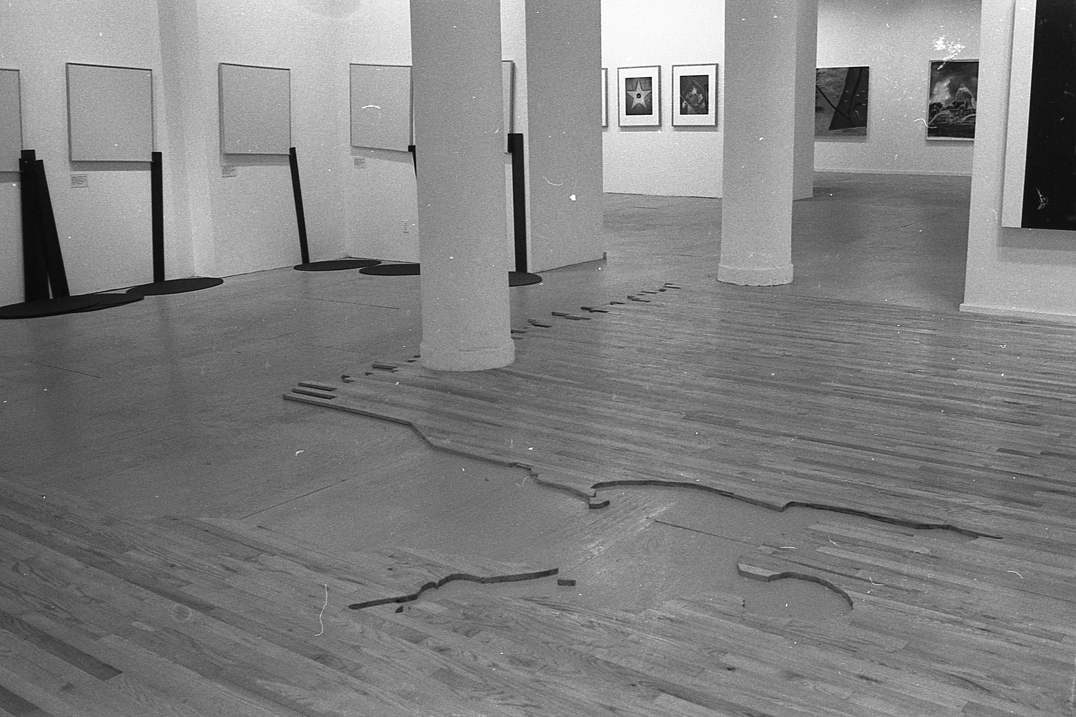 A photograph of a large gallery space with multiple rooms visible.  In the foreground, the floor is covered with flooring, except for a silhouette of a human figure which has been cut out of the flooring, through which the concrete below is visible.