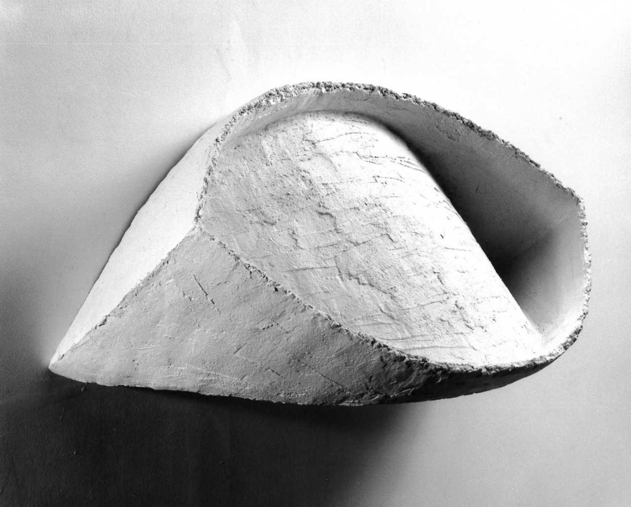 White sculptural form hanging on white gallery wall with stark lighting contrast.