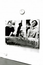 A painting collage with three separate figures represented hanging in a white-walled gallery space with a clock above it right where a finger in painting is pointing.