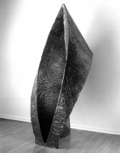A large sculptural abstract black object standing upright on the gallery floor behind a white wall.