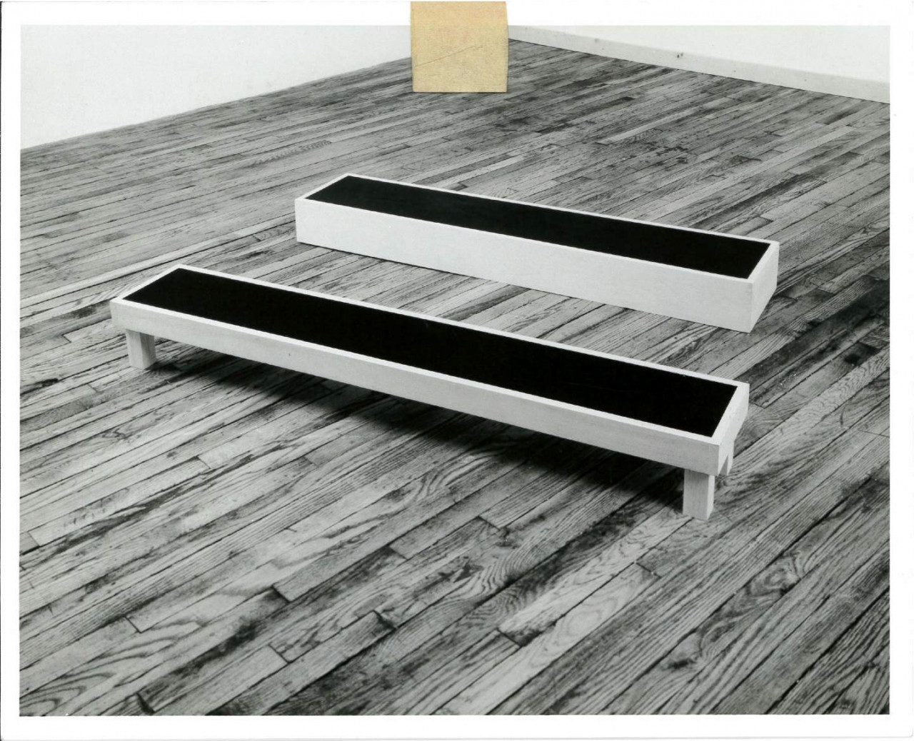 A black and white photograph of gray benches with black on top, the front one with legs the back one without. Tape appears on the top of the image.