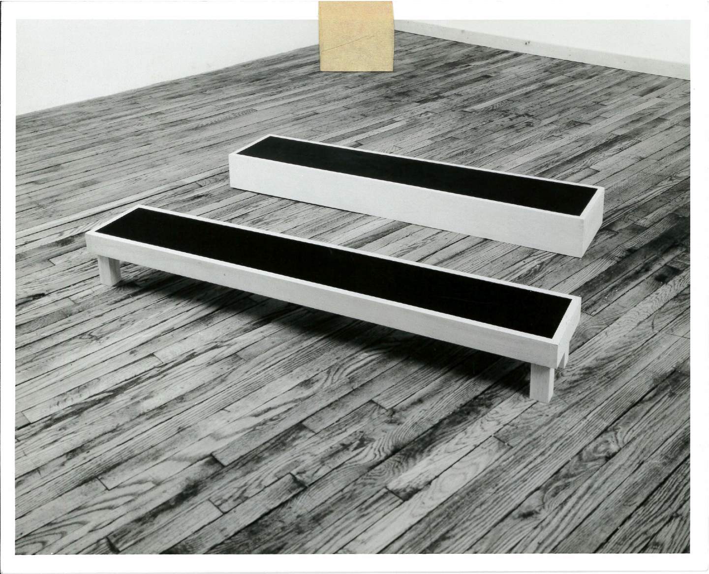 A black and white photograph of gray benches with black on top, the front one with legs the back one without. Tape appears on the top of the image.