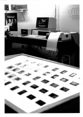 A black and white photograph of a number of unmounted 35mm slides on a lightbox is visible in the foreground with a computer and printer visible in the background in an office setting.