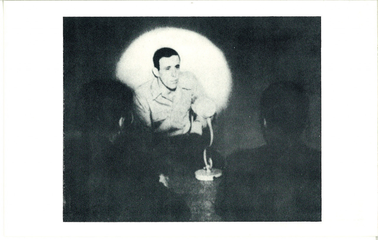 A black-and-white photograph of a man crouching and speaking in front of a spotlight. The outlines of two people in front of the speaker are faintly visible.