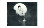 A black-and-white photograph of a man crouching and speaking in front of a spotlight. The outlines of two people in front of the speaker are faintly visible.