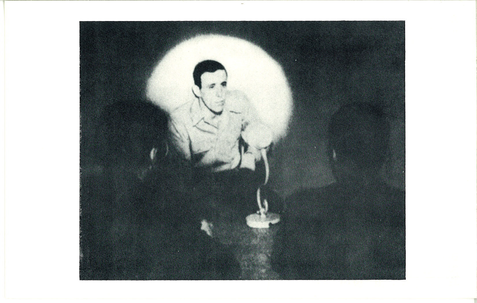 A black-and-white photograph of a man crouching and speaking in front of a spotlight. The outlines of two people in front of the speaker are faintly visible.