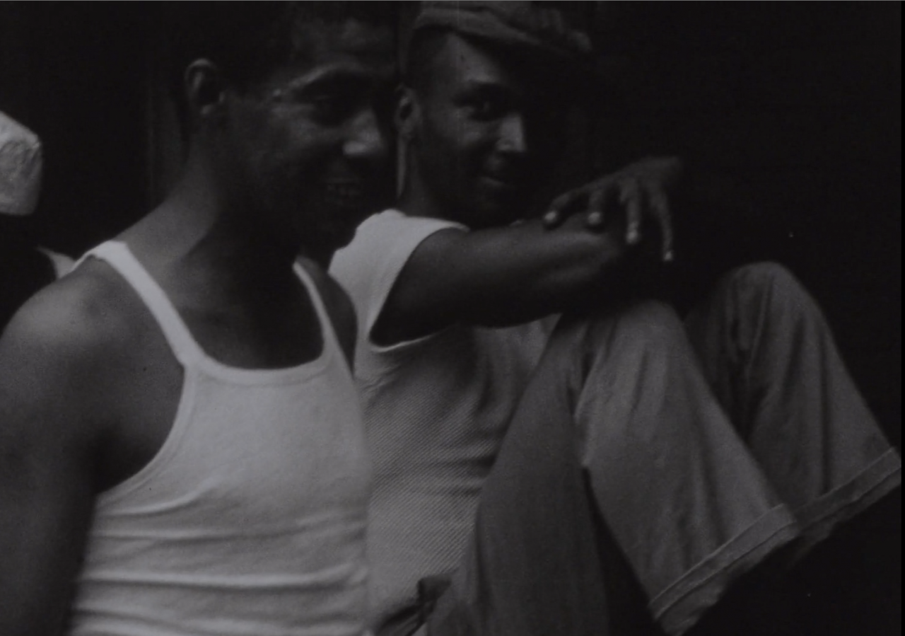 Black-and-white image of two figures, each wearing white shirts. The figure on the right sits with their knees against their chest and looks attentively at the figure to their left, who engages with someone out of the right-side of the frame.