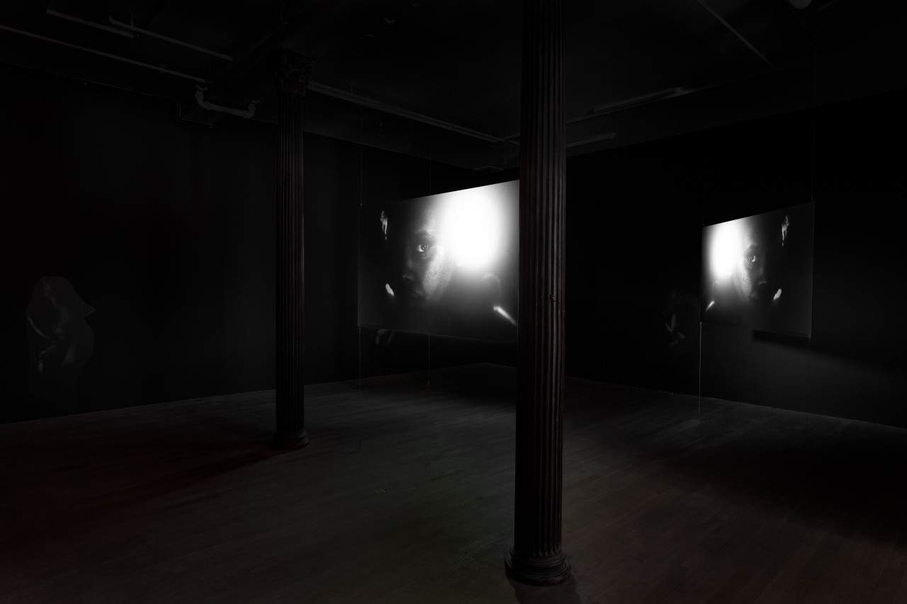Two hanging projector screens are placed in the middle of a darkened gallery room. They both show the same black-and-white video still of a figures face, illuminated on one side by a bright flash of light.