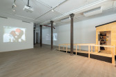 A gallery space with three columns running down the right side. Projected onto a wall on the left is a video still of a woman in a red blanket hunched over. Two projector screens can be seen on the far left wall. In the back is a ramp which leads to the doorway of a wooden camper structure.