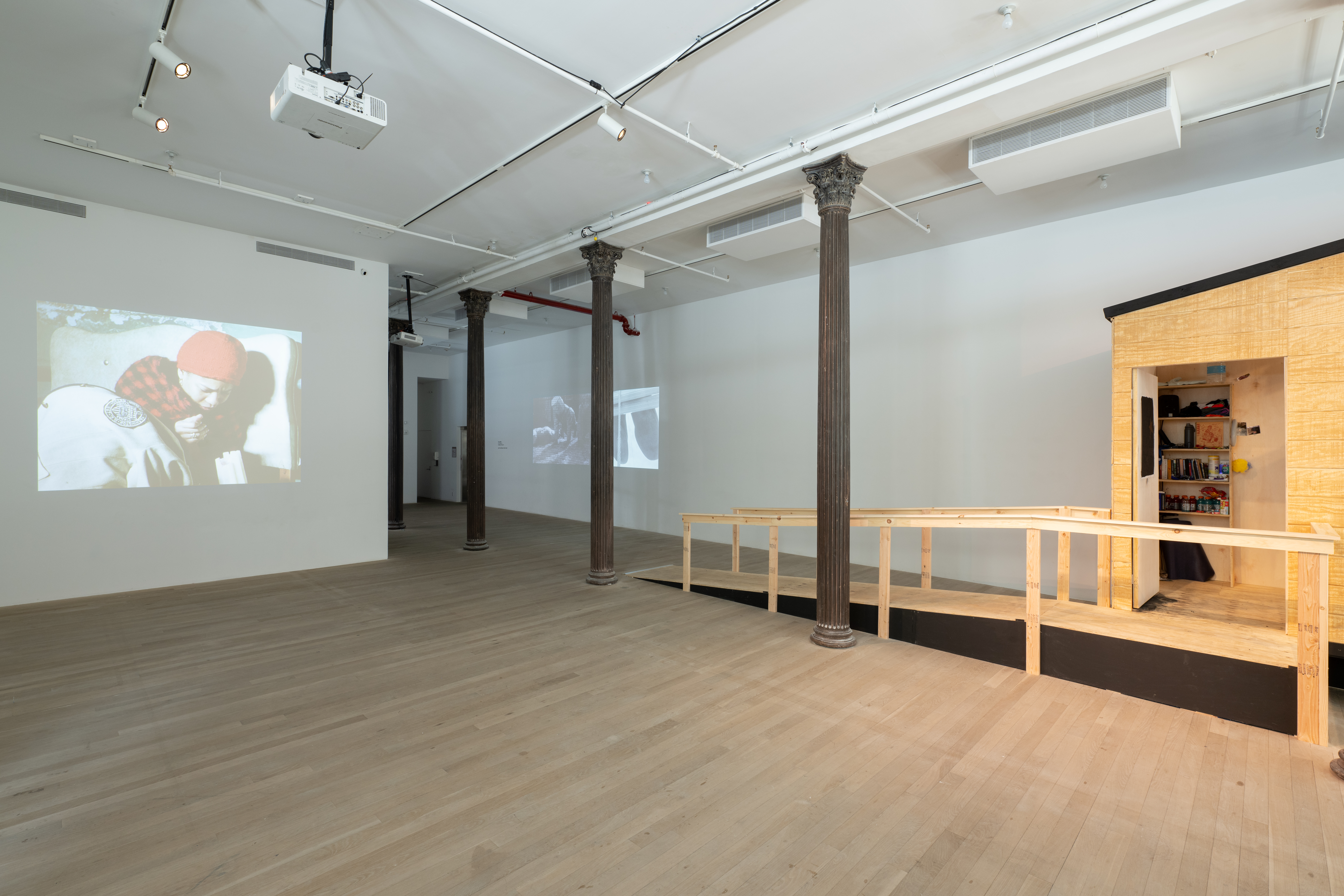 A gallery space with three columns running down the right side. Projected onto a wall on the left is a video still of a woman in a red blanket hunched over. Two projector screens can be seen on the far left wall. In the back is a ramp which leads to the doorway of a wooden camper structure.