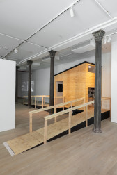 The corner of a gallery space with a wooden camper structure placed there. Two ramps lead to doorways along the front and side of the structure. The edge of a free-standing white wall sits to the left of the camper..