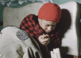 A color film still of an individual curled against a worn beige headboard as they lean into a candle. They don a red beanie and a checkered shawl, covered by a vintage coat emblazoned with a Howard University logo. The immediate background depicts a dilapidated green wall behind the headboard.