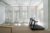 A gallery space with two large black treadmills placed on the right side of the room. Along the back and side are doors with large window panes that show the street outside. The edge of a wooden railing can be seen to the left of the treadmills.