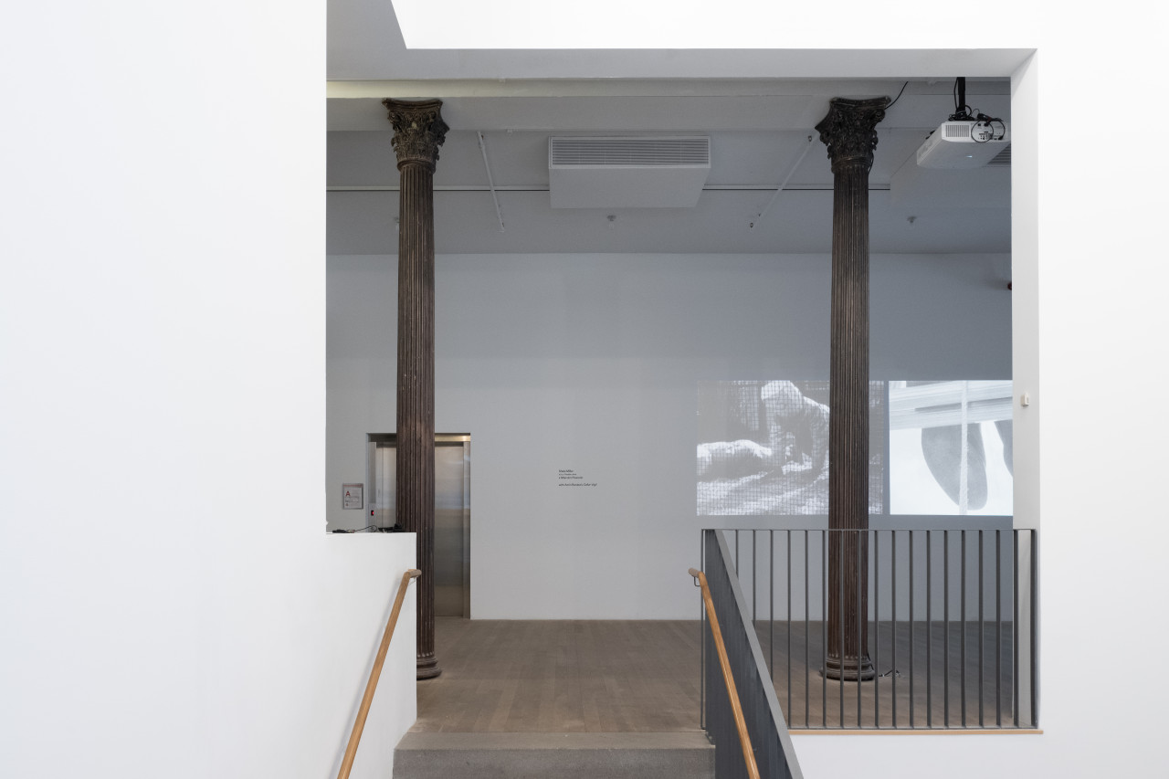 The entrance to a gallery space. Two video stills are projected on the wall facing the entrance. The left projector shows a black-and-white film image of two figures in trench coats with paper bags over their heads doing pushups. On the right is a black-and-white film image of a pair of black shoes hanging over a wooden ledge.