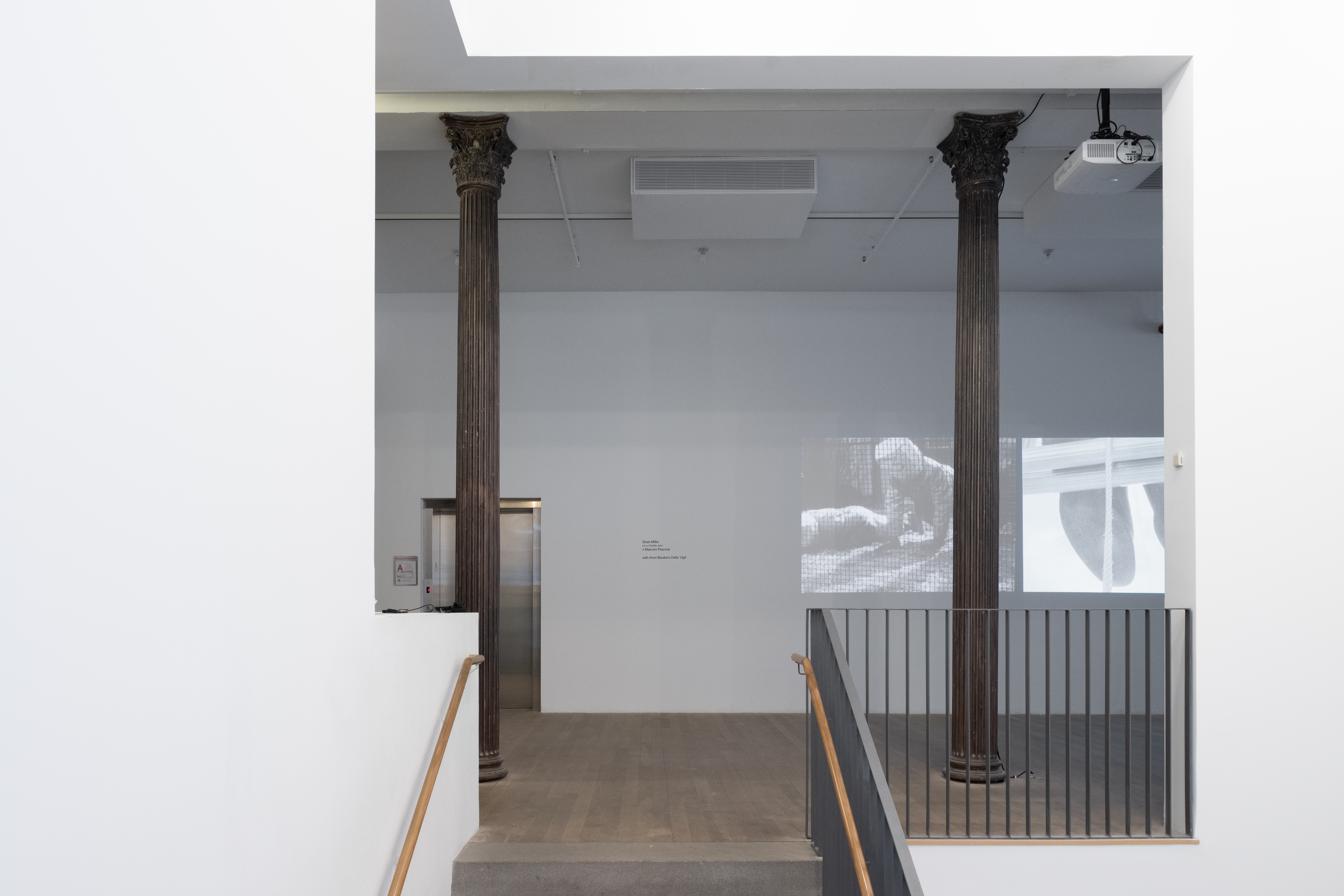 The entrance to a gallery space. Two video stills are projected on the wall facing the entrance. The left projector shows a black-and-white film image of two figures in trench coats with paper bags over their heads doing pushups. On the right is a black-and-white film image of a pair of black shoes hanging over a wooden ledge.