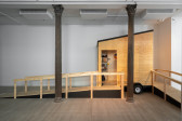 A gallery space with a wooden camper structure situated in the corner of two walls, to the left of the space. Through the open doorway can be seen a small ciruclar window and a shelf with various items placed on it.