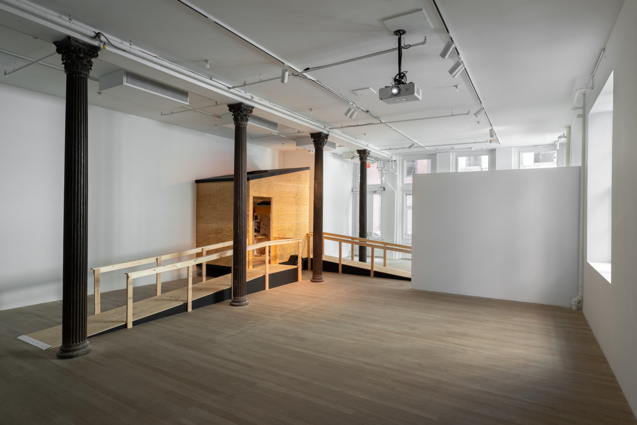 A gallery space with a wooden camper structure placed in the left corner. Two wooden ramps lead to the front and side of the structure. A freedstanding walls juts out from the right wall. A white projector hangs from the ceiling on the right side of the space.