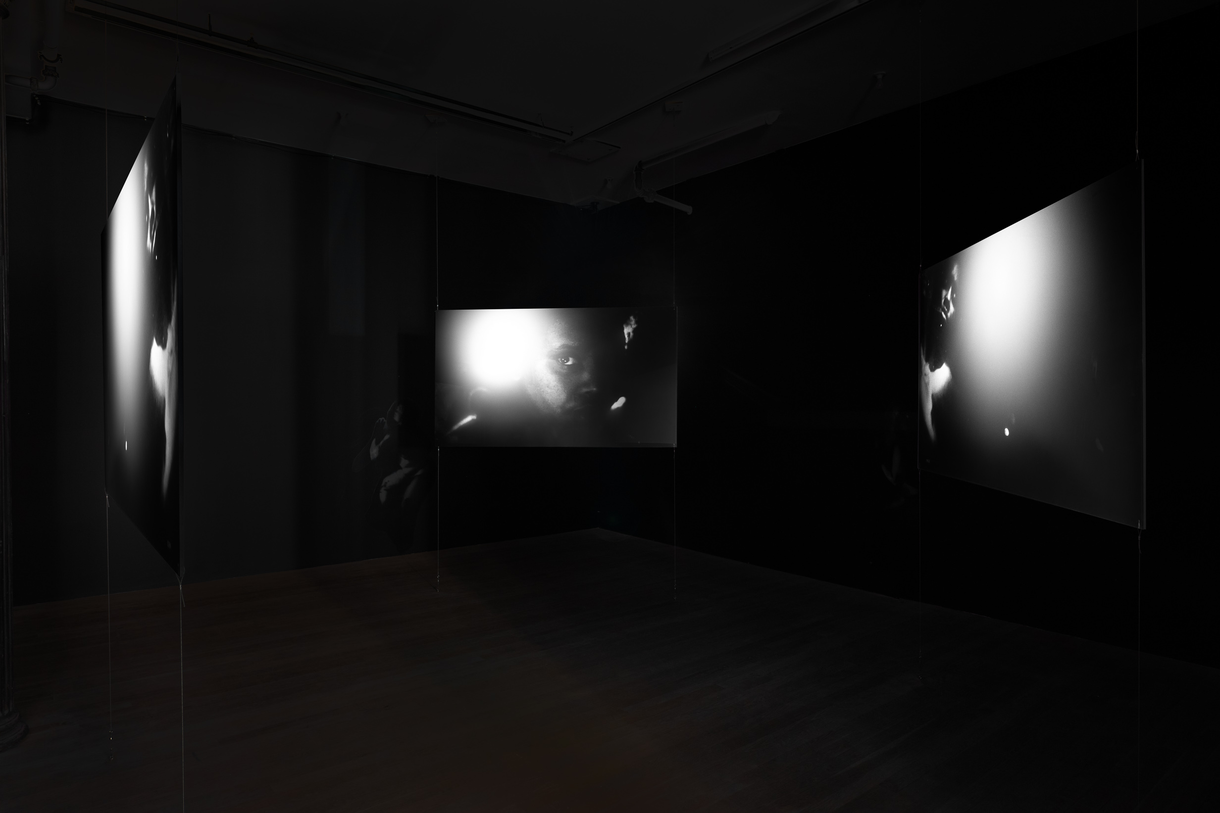 Three hanging projector screens surround the middle of a darkened gallery space on three sides. The screens all show the same black-and-white video still of a figures face, illuminated on one side by a bright flash of light.
