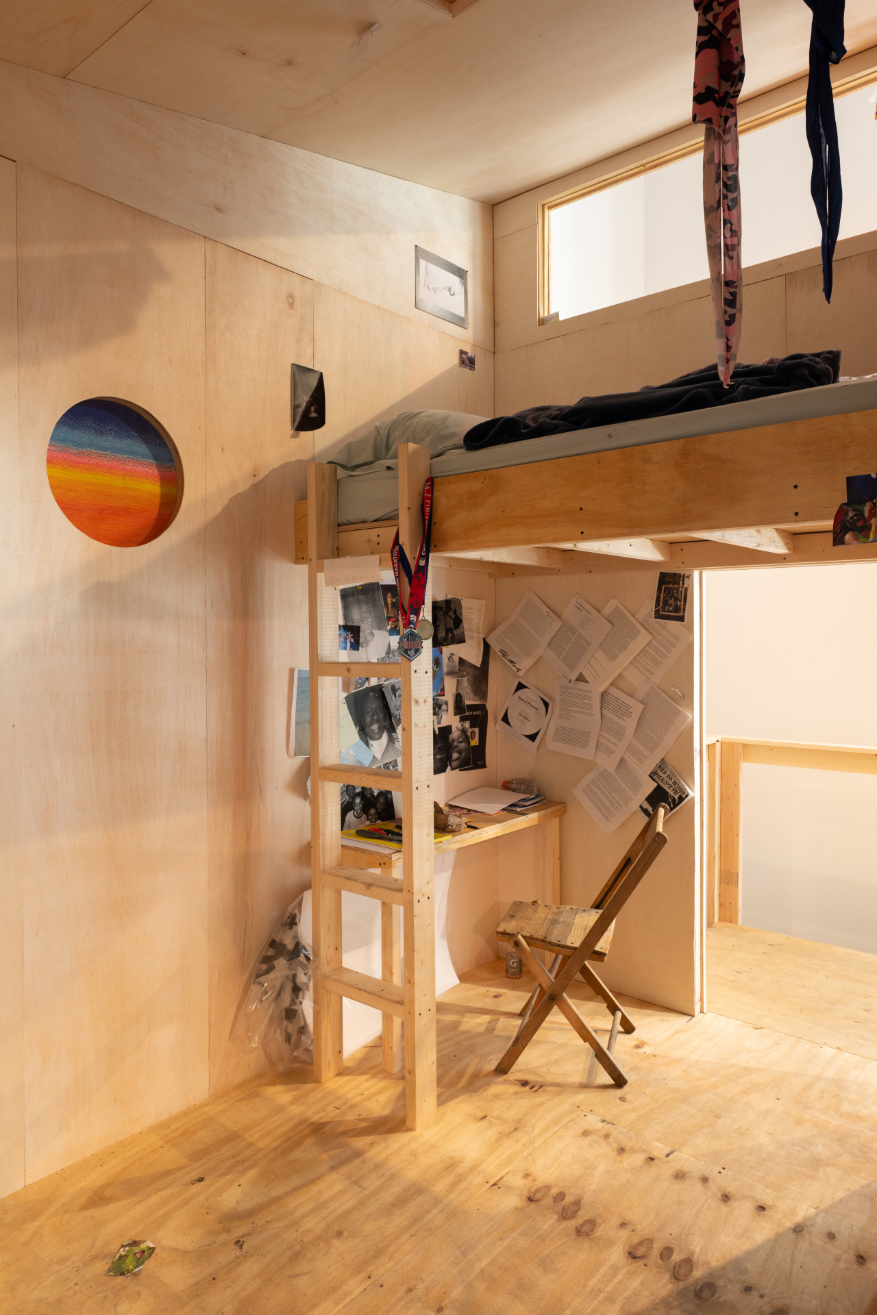 A small wooden room with a circular window depicting a woven gradient of blue to orange. On the right side of the room is a lofted bed with a ladder, under which sits a desk and chair. Various images and documents are pinned to the walls surrounding the desk.