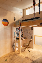 A small wooden room with a circular window depicting a woven gradient of blue to orange. On the right side of the room is a lofted bed with a ladder, under which sits a desk and chair. Various images and documents are pinned to the walls surrounding the desk.