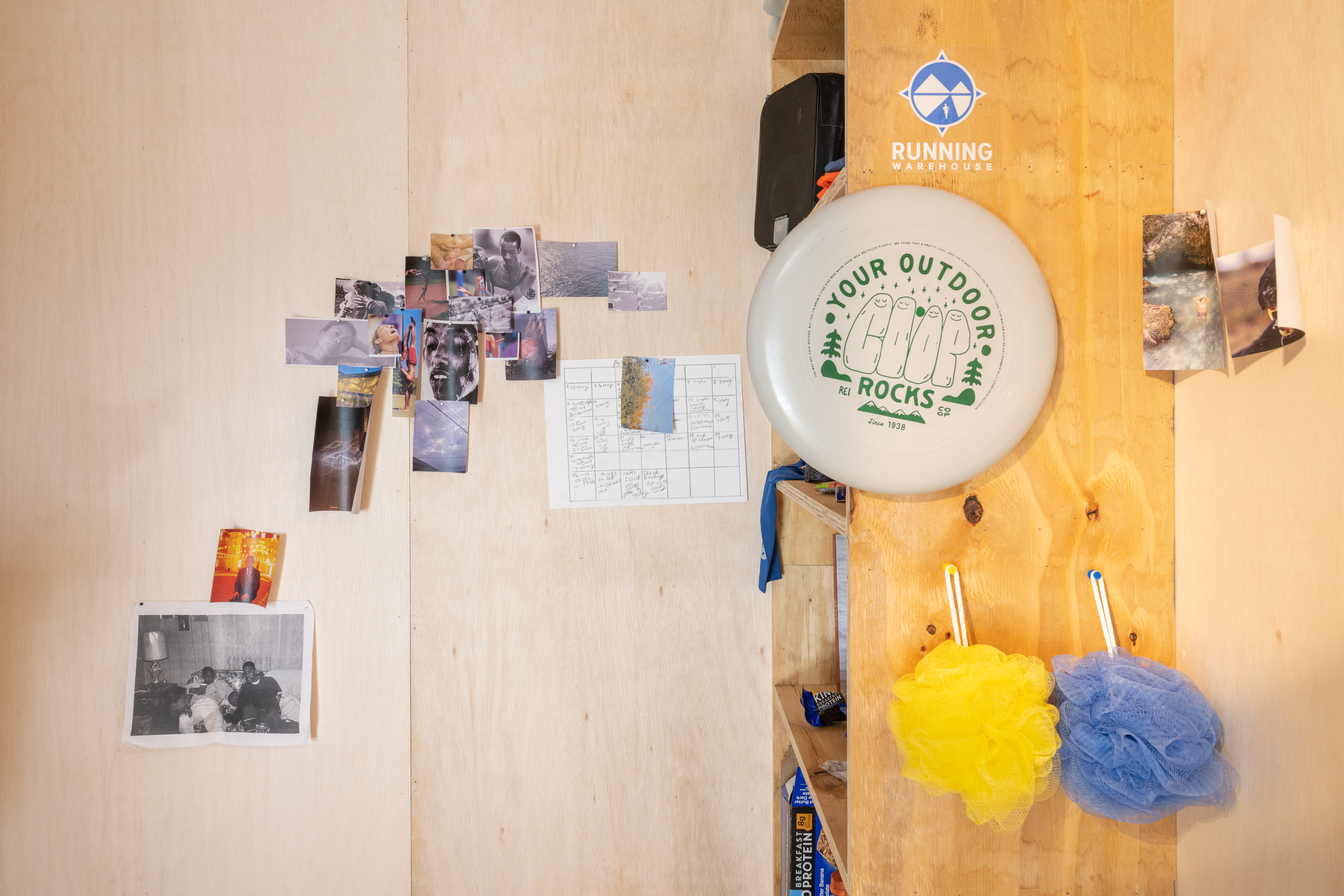 Various photographs of athletes and other personal images are arranged on a light wooden wall to the left. A wooden shelf sits against the right wall, shown in profile. A frisbee and two loofahs hang off the side of the shelf, with other stickers and printed images arranged on the walls.