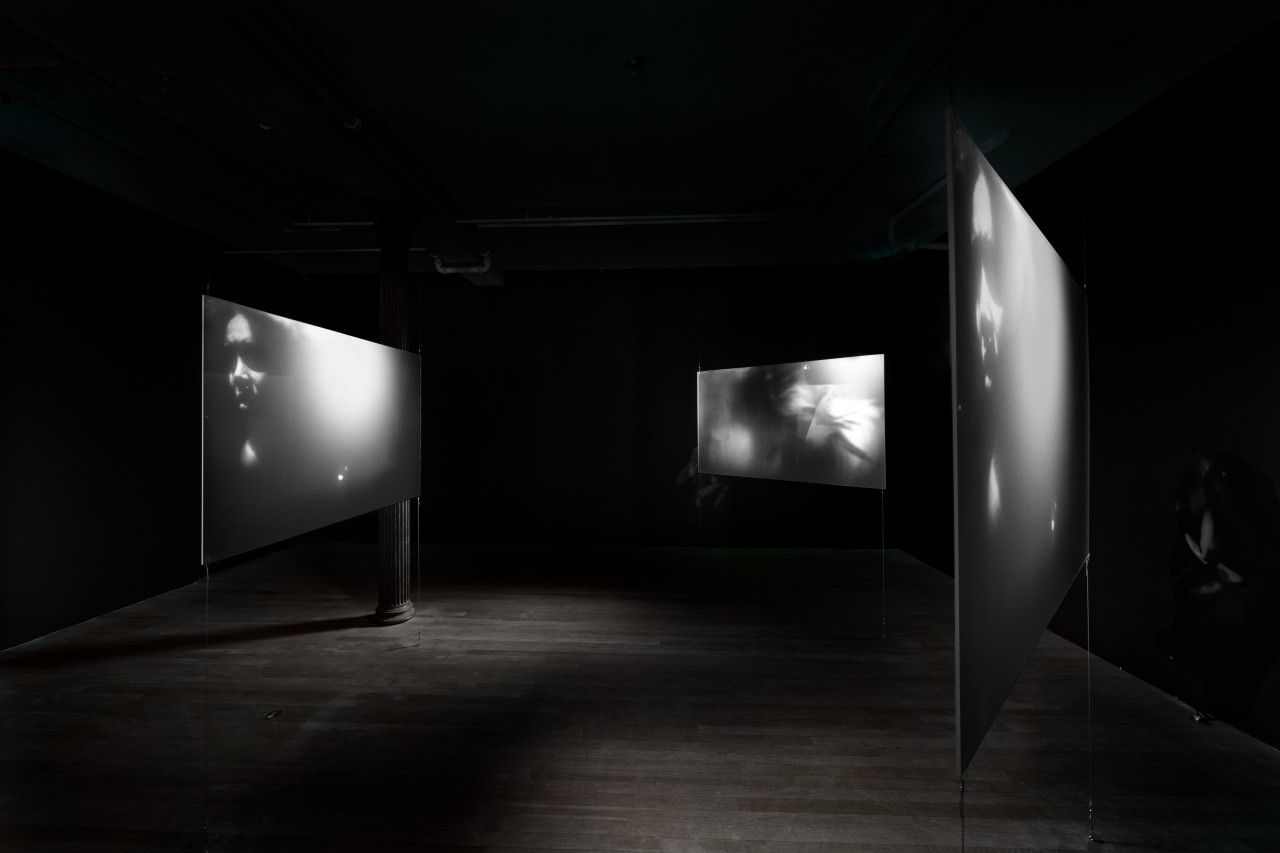 Three hanging projector screens surround the middle of a darkened gallery space. The two screens on the right and left side depict a black-and-white video still of a figures face, illuminated on one side by a bright flash of light. The third screen in the middle shows a black-and-white film still of hazy and ghostly tonal modulations.