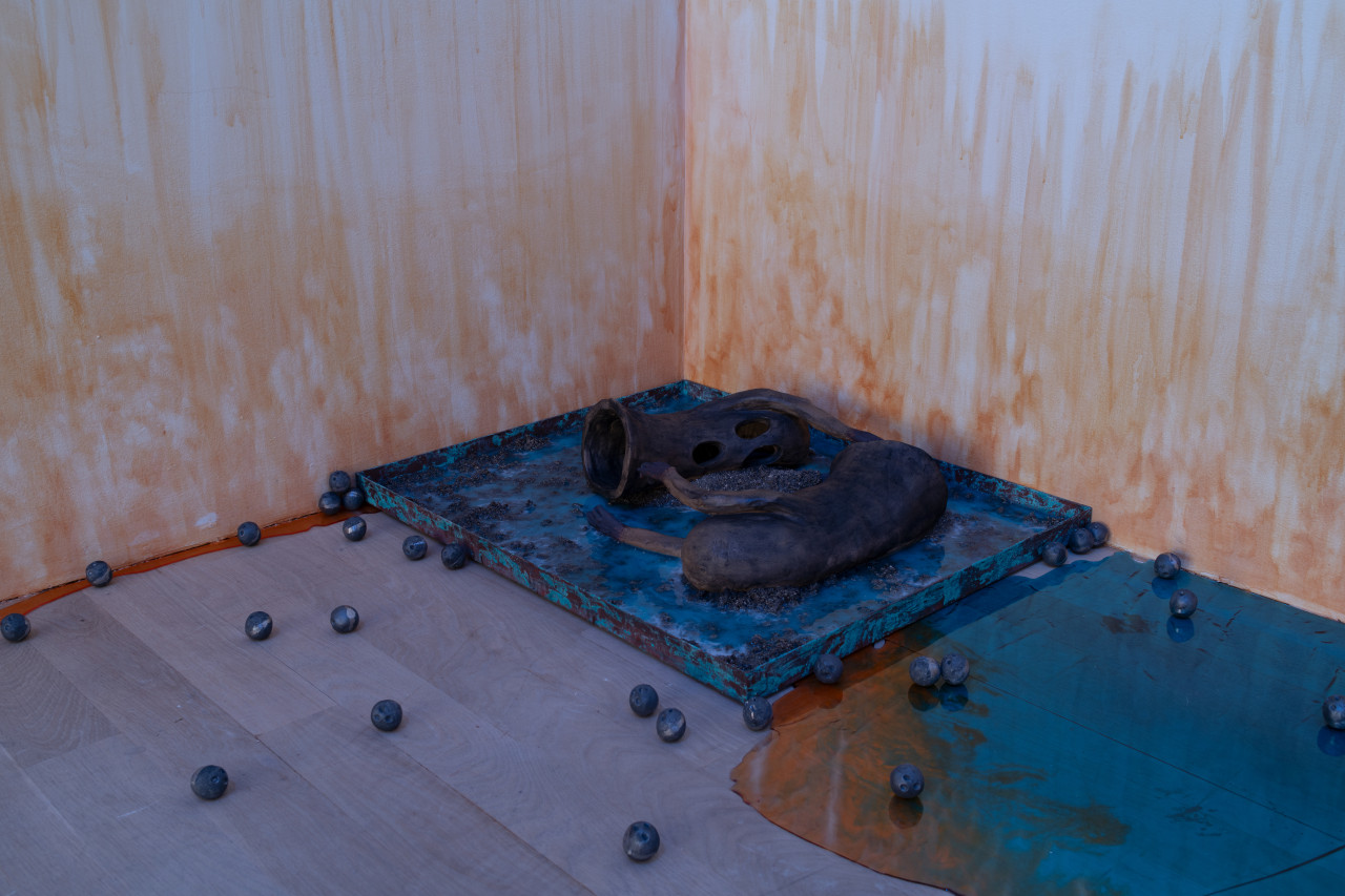 In a corner of the gallery, two oblong ceramic ghosts lay on their sides on top of a rusty blue square with small piles of charcoal. A shiny blue and orange pool of resin appears to leak out along the edges of the wall and numerous small metal balls are scattered around. On the wall, an orange stain appears to emerge from the floor.