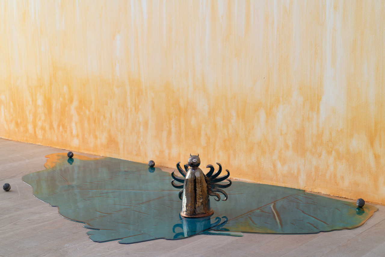 A small cylindrical ceramic figure, with curly filamented wings and a round head studded with horns and cowrie shell eyes, sits atop a reflective blue pool of resin with orange tints at the edges. Four small metal balls are scattered around and, on the wall behind, an orange stain appears to emerge from the floor.