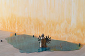 A small cylindrical ceramic figure, with curly filamented wings and a round head studded with horns and cowrie shell eyes, sits atop a reflective blue pool of resin with orange tints at the edges. Four small metal balls are scattered around and, on the wall behind, an orange stain appears to emerge from the floor.