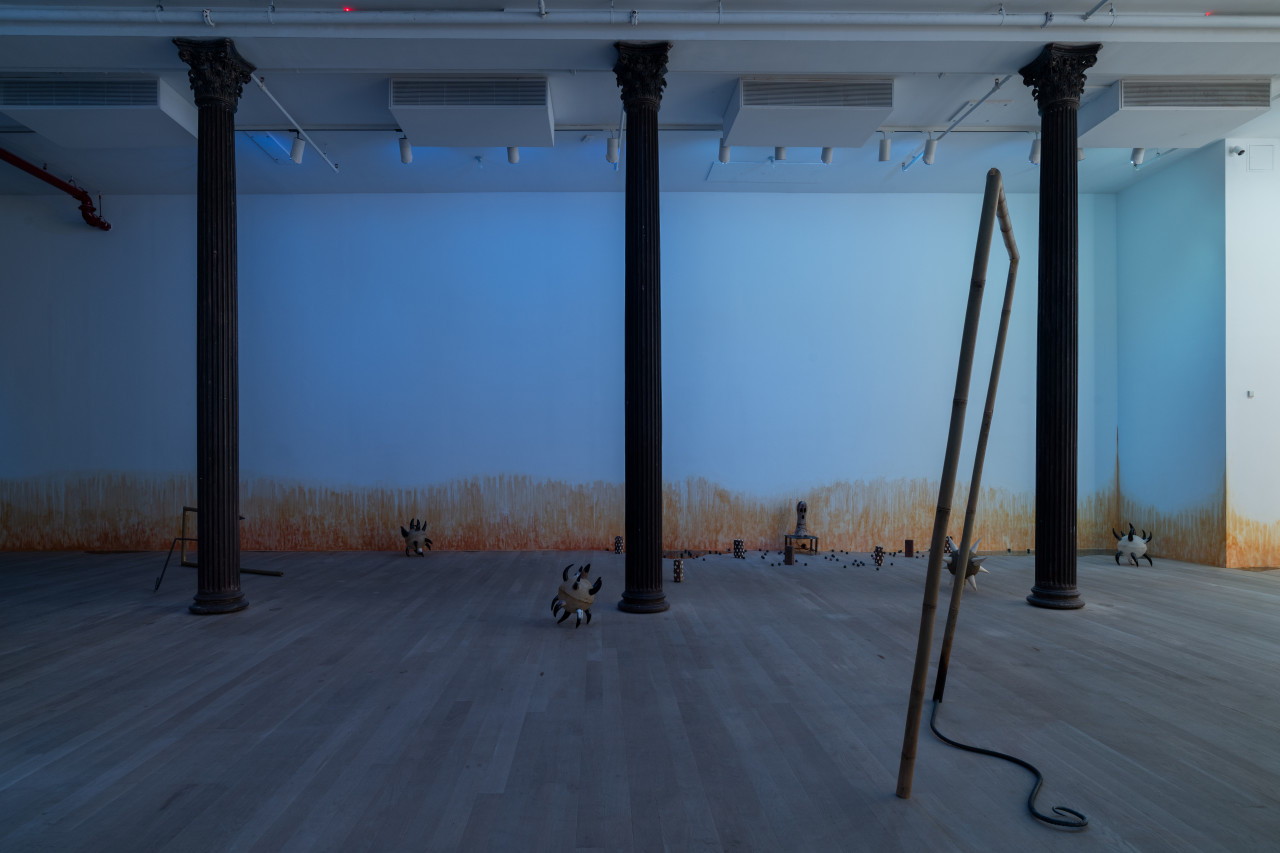 Three columns are centered in a blue-lit gallery space. Various sculptures are arranged on the floor, including morning stars with cow horn spikes; two bamboo and rebar spears shaped into rectangles; bricks wrapped in cowrie shells; a small oblong ceramic figure on a short stool; resin pools; and ceramic balls. On the wall, an orange stain appears to emerge from the floor.