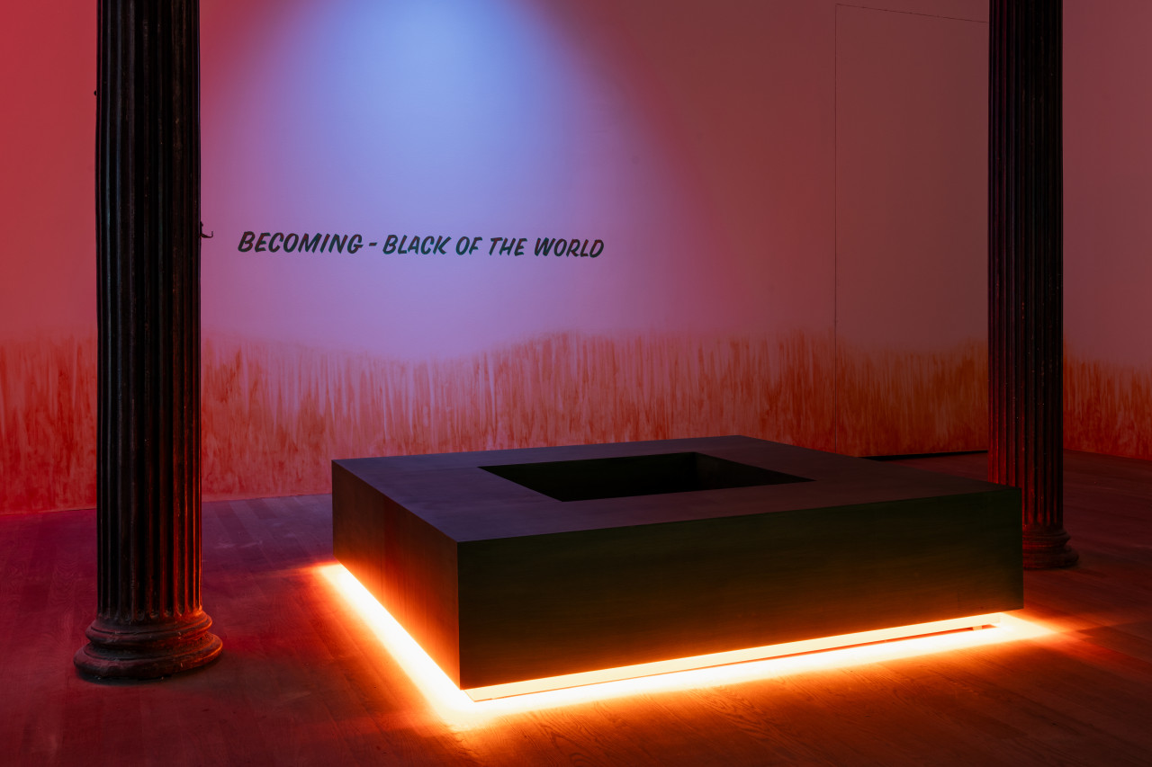 Between two columns in a darkened room, a square-shaped bench radiates bright orange light from below. On the wall, an orange stain appears to emerge from the floor while words "BECOMING - BLACK OF THE WORLD" are illuminated by a blue light.