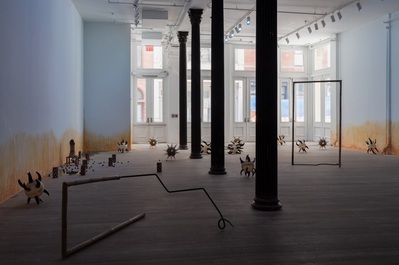 A sunlit gallery space is bisected by four columns through the middle. Various sculptures are arranged on the floor, including morning stars with cow horn spikes; two bamboo and rebar spears shaped into rectangles; bricks wrapped in cowrie shells; a small oblong ceramic figure on a short stool; resin pools; and ceramic balls. On the wall, an orange stain appears to emerge from the floor.
