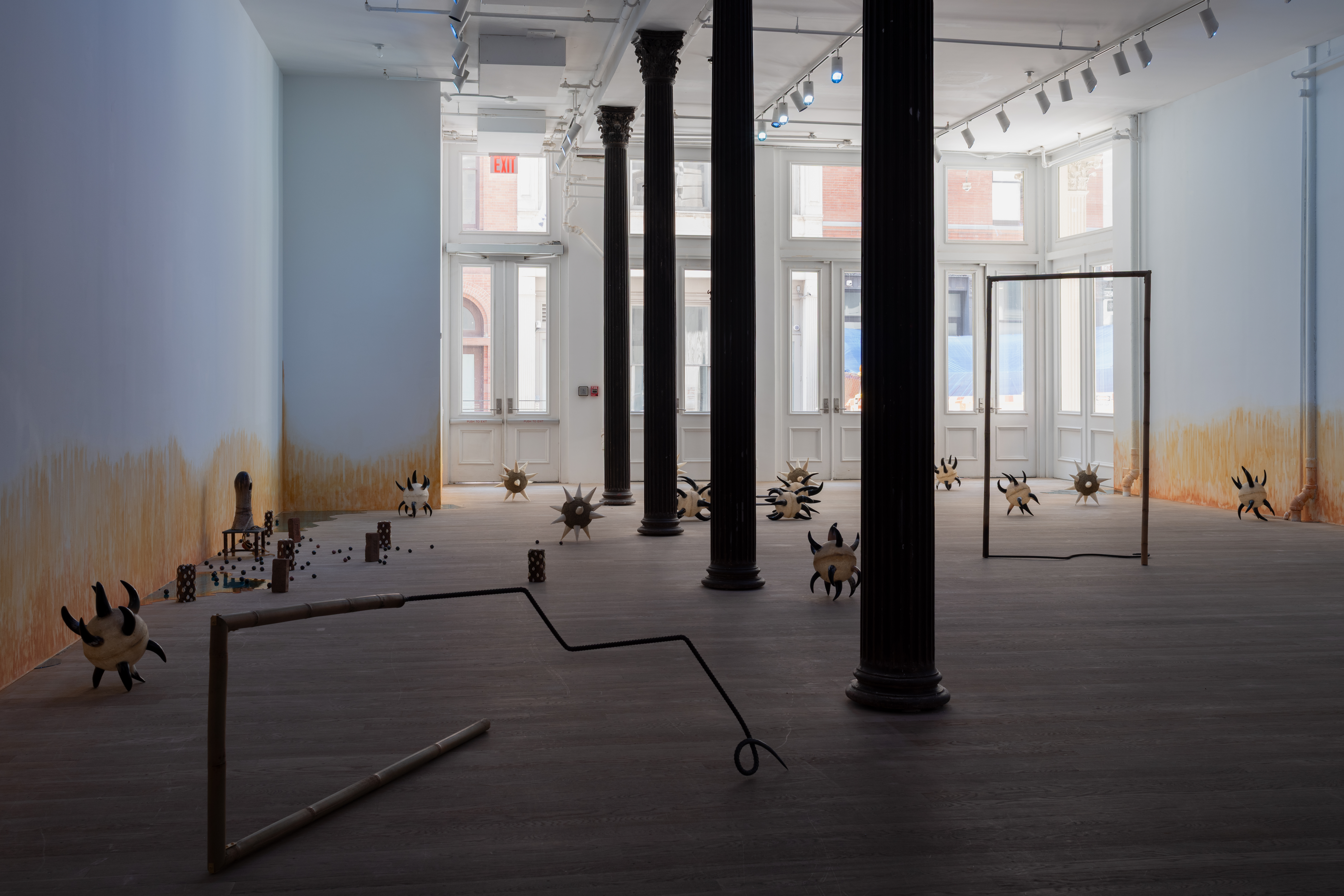 A sunlit gallery space is bisected by four columns through the middle. Various sculptures are arranged on the floor, including morning stars with cow horn spikes; two bamboo and rebar spears shaped into rectangles; bricks wrapped in cowrie shells; a small oblong ceramic figure on a short stool; resin pools; and ceramic balls. On the wall, an orange stain appears to emerge from the floor.