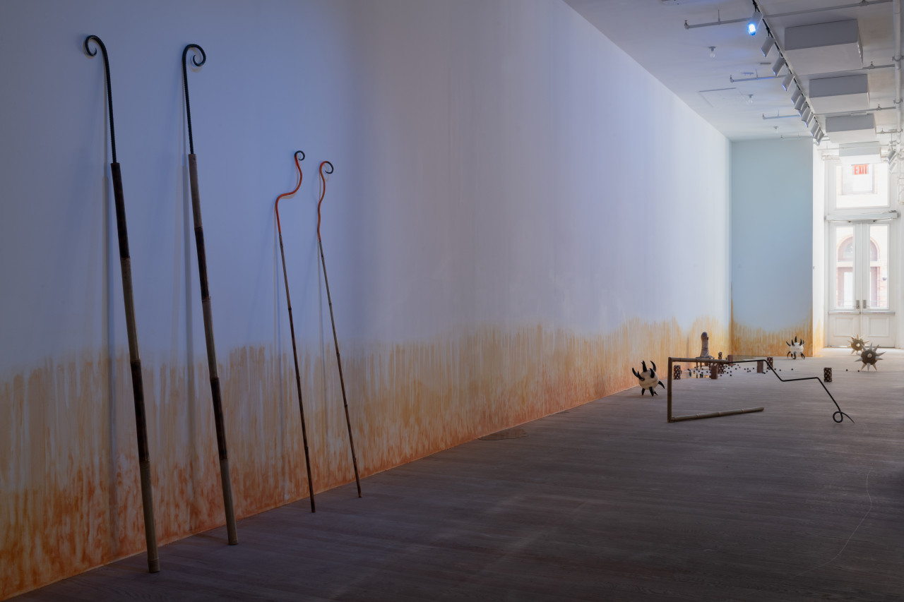 In a blue-lit gallery space, two pairs of bamboo and rebar spears are curled at the tips and leant against the wall. To the right, various sculptures are arranged on the floor, including morning stars with cow horn spikes; a bamboo and rebar spear shaped into a rectangle; bricks wrapped in cowrie shells; a small oblong ceramic figure on a short stool; resin pools; and ceramic balls. On the wall, an orange stain appears to emerge from the floor.