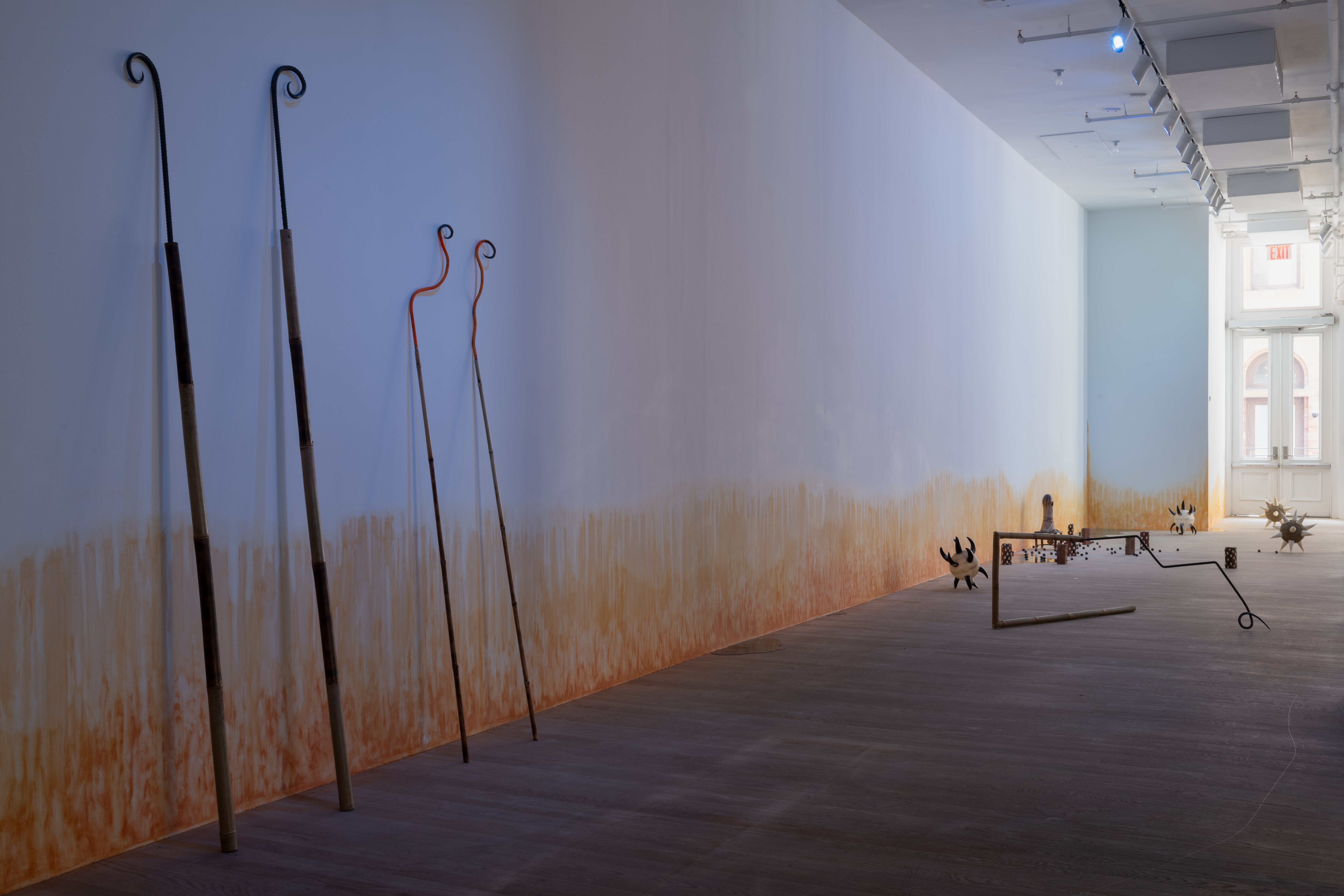 In a blue-lit gallery space, two pairs of bamboo and rebar spears are curled at the tips and leant against the wall. To the right, various sculptures are arranged on the floor, including morning stars with cow horn spikes; a bamboo and rebar spear shaped into a rectangle; bricks wrapped in cowrie shells; a small oblong ceramic figure on a short stool; resin pools; and ceramic balls. On the wall, an orange stain appears to emerge from the floor.