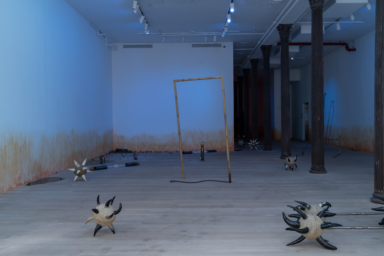 A blue-lit gallery space is filled with various sculptures arranged on the floor, including including morning stars with cow horn spikes; two bamboo and rebar spears shaped into rectangles; bricks wrapped in cowrie shells; sugar cane rollers; resin pools; and ceramic balls. On the wall, an orange stain appears to emerge from the floor.