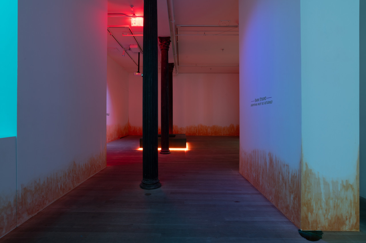 In a darkened space lit by blue light, a wall to the right reads "SHANI STRAND / EVERYTHING MUST BE RETURNED." Further into the room, a square-shaped bench between two columns radiates bright orange light from below. On the wall, an orange stain appears to emerge from the floor.