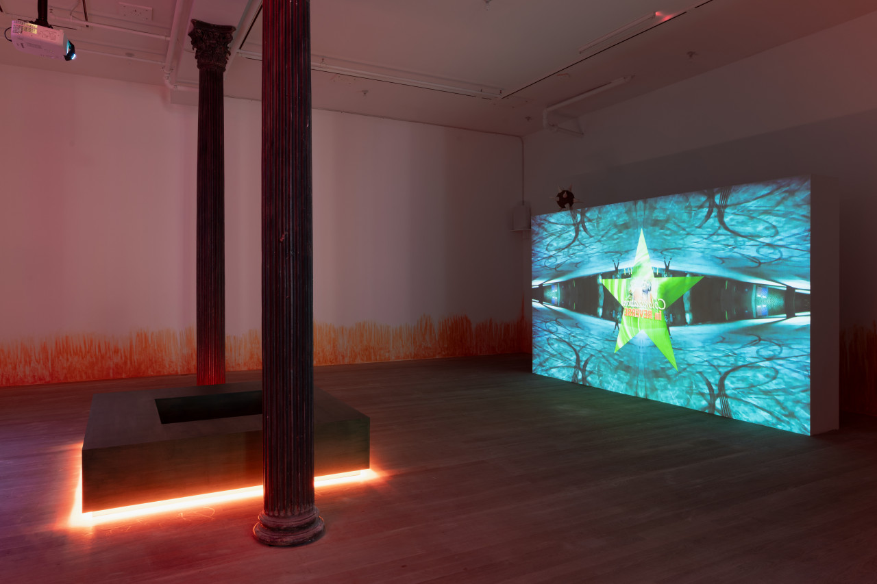 On the left between two columns in a darkened room, a square-shaped bench radiates bright orange light from below. On the right, a large projection on the wall shows a green star with the text "Colonization / in REVERSE" superimposed on top of an image of a figure standing in a parking lot, which has been reflected four times.